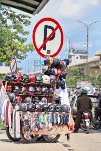 A vendor is displaying helmets and caps on handcart on footpath under a 'No Parking' sign along Murree Road.