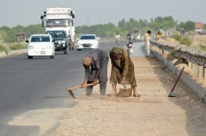 Labourers engaged in leveling and compacting gravel using a road roller as part of ongoing road construction work to enhance transportation facilities near Head Muhammad Wala.