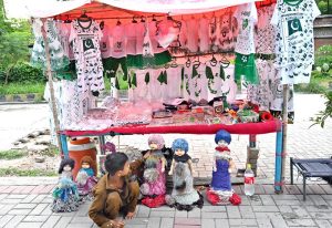 A young vendor arranging and displaying national flag color children dresses and other items related to Independence Day celebrations at his roadside setup.