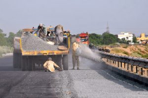 Labourers engaged in leveling and compacting gravel using a road roller as part of ongoing road construction work to enhance transportation facilities near Head Muhammad Wala.
