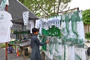 A young vendor arranging and displaying national flag color children dresses and other items related to Independence Day celebrations at his roadside setup.