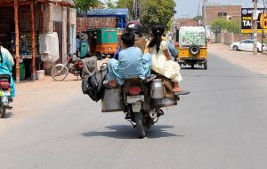 An overloaded motorcycle carries two children and multiple milk containers on a busy road, posing a risk to the children’s lives.