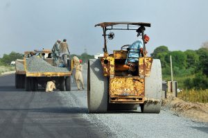 Labourers engaged in leveling and compacting gravel using a road roller as part of ongoing road construction work to enhance transportation facilities near Head Muhammad Wala.