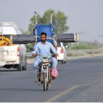 A man riding a motorcycle loaded with a chair and curtain (chic) tied at the back, navigating through traffic on a busy road, depicting unsafe transportation methods, overloading, and violation of road safety regulations