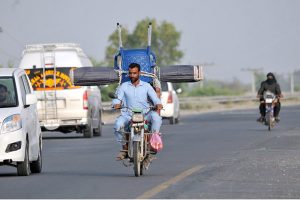 A man riding a motorcycle loaded with a chair and curtain (chic) tied at the back, navigating through traffic on a busy road, depicting unsafe transportation methods, overloading, and violation of road safety regulations