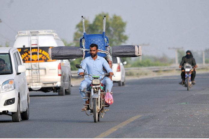 A man riding a motorcycle loaded with a chair and curtain (chic) tied at the back, navigating through traffic on a busy road, depicting unsafe transportation methods, overloading, and violation of road safety regulations