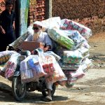 A view of daily toil as an elderly motorcyclist balances a stack of chips and snake packets on his motorcycle during a delivery in Latifabad