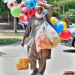 An elderly vendor carrying colorful footballs and eatables to attract children in a public place.