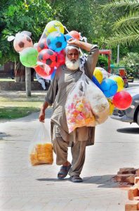 An elderly vendor carrying colorful footballs and eatables to attract children in a public place.