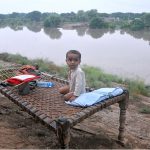 A child sitting on a traditional bed (charpai) waiting for food after being displaced from his home due to heavy floods, with submerged houses visible in the background