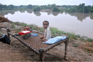 A child sitting on a traditional bed (charpai) waiting for food after being displaced from his home due to heavy floods, with submerged houses visible in the background