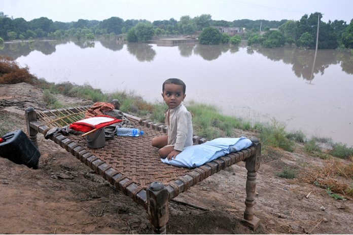 A child sitting on a traditional bed (charpai) waiting for food after being displaced from his home due to heavy floods, with submerged houses visible in the background