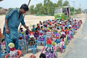 A roadside vendor neatly arranges colorful handmade dolls in the rows on plastic chairs to attract buyers along a busy road.