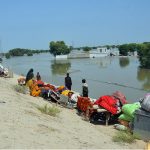 Flood-affected families sit on higher ground with their salvaged belongings, waiting for rescue teams after rising floodwaters swallowed their village, displacing thousands and destroying homes, crops, and livestock.