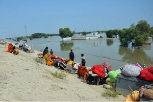 Flood-affected families sit on higher ground with their salvaged belongings, waiting for rescue teams after rising floodwaters swallowed their village, displacing thousands and destroying homes, crops, and livestock.