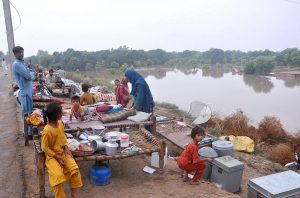 A child sitting on a traditional bed (charpai) waiting for food after being displaced from his home due to heavy floods, with submerged houses visible in the background