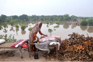 A child sitting on a traditional bed (charpai) waiting for food after being displaced from his home due to heavy floods, with submerged houses visible in the background