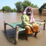 A villager sits on a charpoy in knee-deep water as floodwater submerge his home and village