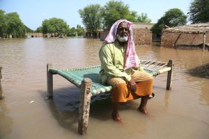 A villager sits on a charpoy in knee-deep water as floodwater submerge his home and village