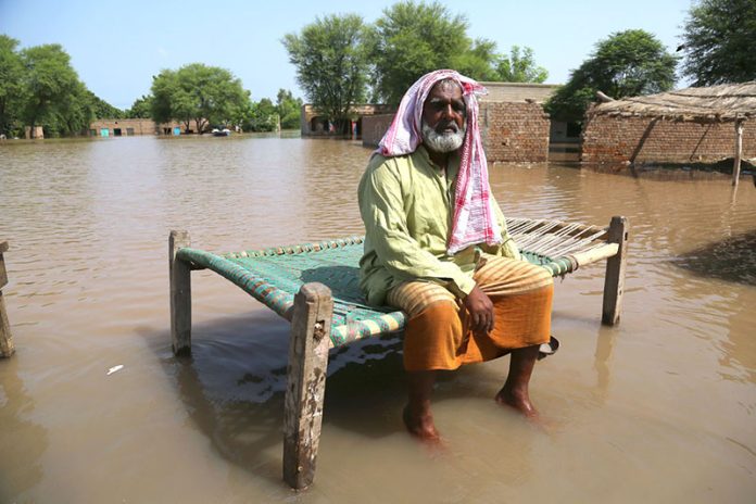 A villager sits on a charpoy in knee-deep water as floodwater submerge his home and village