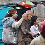 A family rides a motorcycle as a woman shields the rider with a small umbrella to protect them from the rain in the city