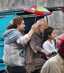 A family rides a motorcycle as a woman shields the rider with a small umbrella to protect them from the rain in the city