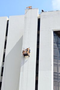 A painter busy painting the exterior wall of the State Bank building while working on a suspended platform.