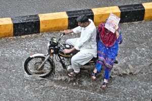 A couple rides a motorcycle through accumulated rainwater during a rain shower in the city.