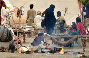 A nomadic woman cooks over an open flame as families rest on cots in a makeshift shelter, demonstrating the resilience of displaced people in difficult circumstances