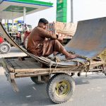 A young labourer sits on large iron sheets being transported on a motorcycle loader near Nag Shah Chowk