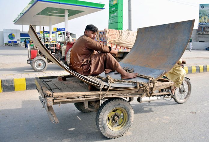 A young labourer sits on large iron sheets being transported on a motorcycle loader near Nag Shah Chowk