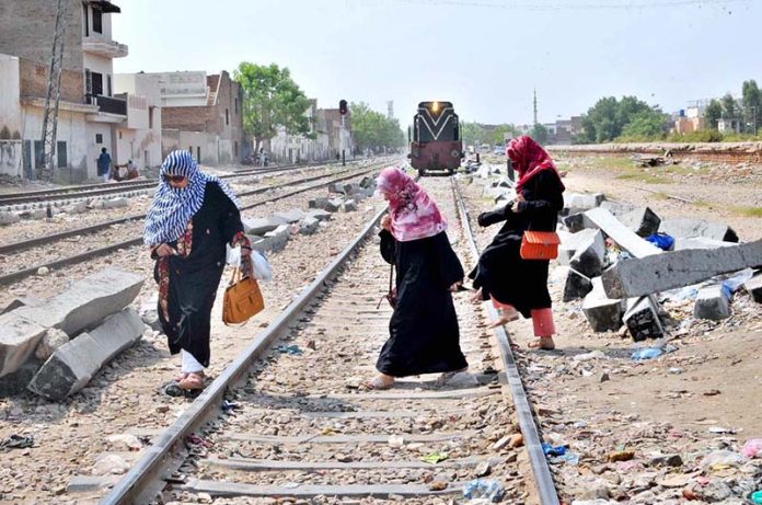 Women cross railway tracks as a train approaches in the background, highlighting unsafe pedestrian practices near railways in urban areas
