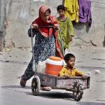 A gypsy girl pushes a cart carrying a young boy and water containers along the roadside in a low-income area, highlighting daily struggles for basic necessities