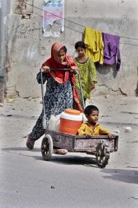 A gypsy girl pushes a cart carrying a young boy and water containers along the roadside in a low-income area, highlighting daily struggles for basic necessities