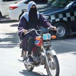 A veiled woman rides a motorcycle without a helmet on a busy road in the city.