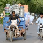 A three-wheeler driver dangerously overloads his vehicle with goods while driving on a city road.