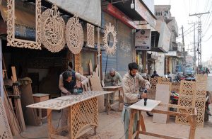 Workers carve and shape decorative wooden panels at a roadside workshop.