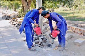 Two laborers operate jackhammers to break concrete on the roadside during ongoing construction.