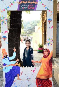 A young girl poses for a photo inside a colorful festival-themed frame during the ten-day annual folk festival “Lok Mela 2025” at Lok Virsa.