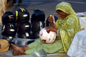 A female street vendor adjusts a helmet while waiting for customers along a roadside in the city.