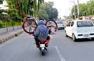A woman holding a bicycle while sitting on the rear seat of a motorcycle on a city road.