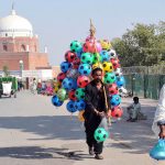 A street vendor walks along the roadside carrying colorful inflatable footballs for sale near the historic Shrine of Hazrat Bahauddin Zakariya.