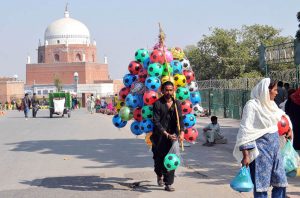 A street vendor walks along the roadside carrying colorful inflatable footballs for sale near the historic Shrine of Hazrat Bahauddin Zakariya.