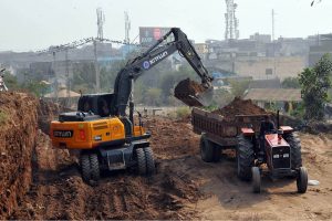 Heavy machinery operates as an excavator lifts soil and loads it onto a tractor-trolley during development and earth-cutting work at Qila Qasim Bagh