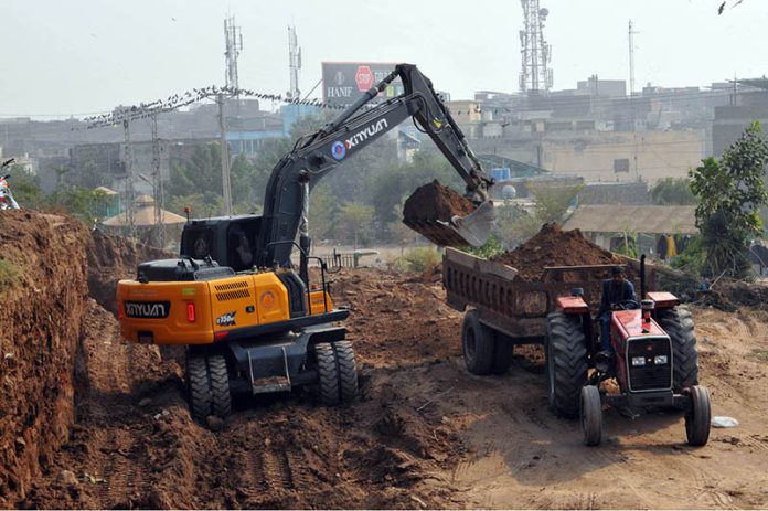 Heavy machinery operates as an excavator lifts soil and loads it onto a tractor-trolley during development and earth-cutting work at Qila Qasim Bagh