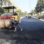 Workers operate a road roller to level and carpet the road with compact gravel on a damaged section as part of ongoing construction work aimed at improving transportation facilities near the IJP Service Road