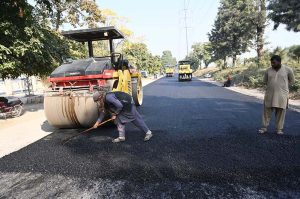 Workers operate a road roller to level and carpet the road with compact gravel on a damaged section as part of ongoing construction work aimed at improving transportation facilities near the IJP Service Road