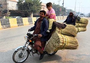 An overloaded motorcyclist is carrying a broom and carrying his child sits on the broom in a dangerous manner, posing a serious risk of accident on Qambar Road.