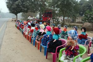 Colorful handmade dolls are displayed on small plastic chairs along a roadside for sale to attract customers.