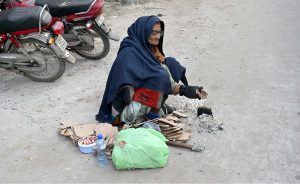 Amid chilly weather, an elderly beggar woman seeks warmth near a roadside fire.